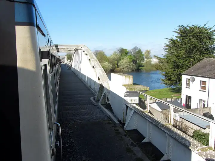 The white iron arch of Athlone Railway Bridge seen from a train crossing it, with the Shannon and its tree-lined banks below on a sunny day.