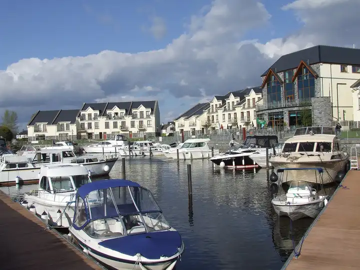 Motor cruisers fill the marina beside the Leitrim Marina Hotel and its apartments, with wooden pontoons in the foreground under a blue sky with clouds.