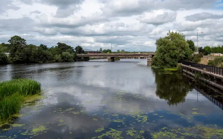 The River Bann at Toome flows wide and calm under a low road bridge, with trees on both banks, a railed walkway on the right and patches of weed on the water.