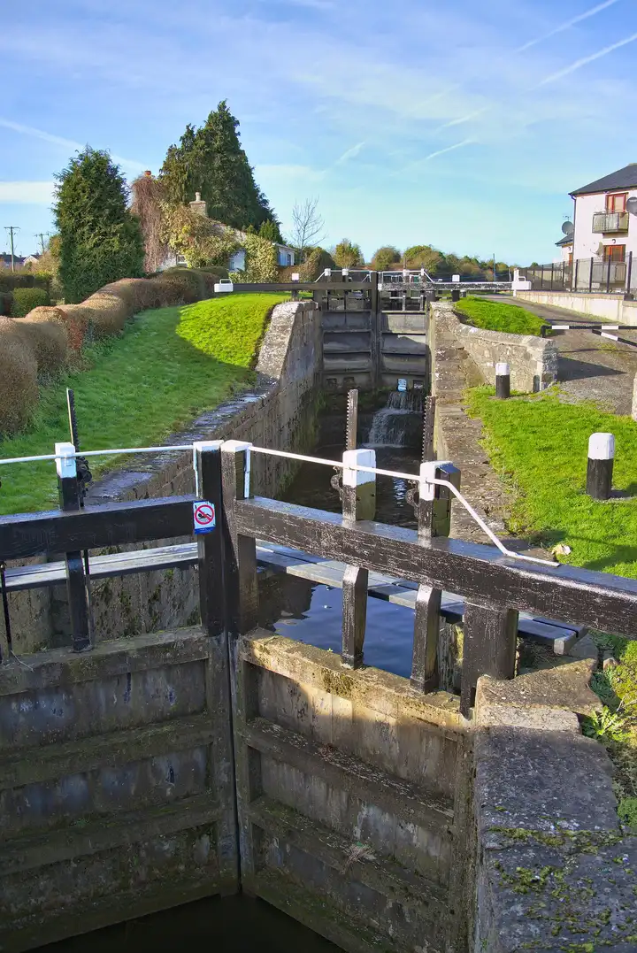 Blick über geschlossene Schleusentore in die doppelte Kammer der 16. Schleuse in Kilcock, wo an einem sonnigen Tag Wasser zwischen grasbewachsenen Ufern aus den oberen Toren läuft.