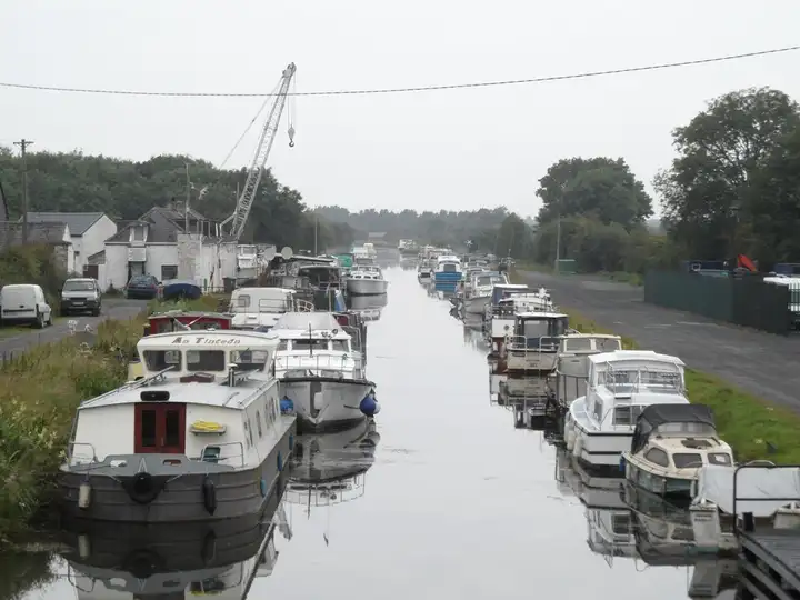 Cruisers and barges moored nose to tail along both banks of the canal at Lowtown, with a boatyard crane and white buildings on the left.