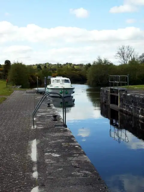 Ein weißes Motorboot in der Schleuse Skelan (Schleuse 3), entlang des steinernen Schleusenrands gesehen, rechts die Schleusentore, dahinter Bäume am Ufer.