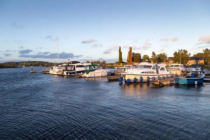 Motor cruisers and a yacht moored at the floating jetties of Dromineer harbour in evening light, with the ruined castle behind.