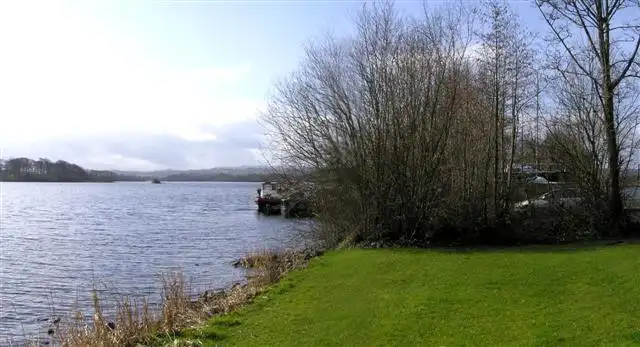 Ein Boot liegt an einem kleinen Steg neben kahlen Bäumen in der Goblusk Bay beim Lough Erne Yacht Club, davor ein grüner Rasen und ein schilfiges Ufer.
