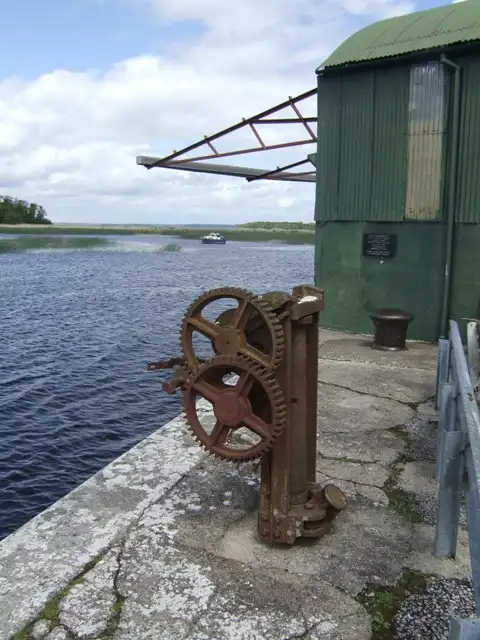 Een oude, verroeste kraanlier op de stenen kade van Kilgarvan naast een groene golfplaten loods, met het meer en een bootje erachter.