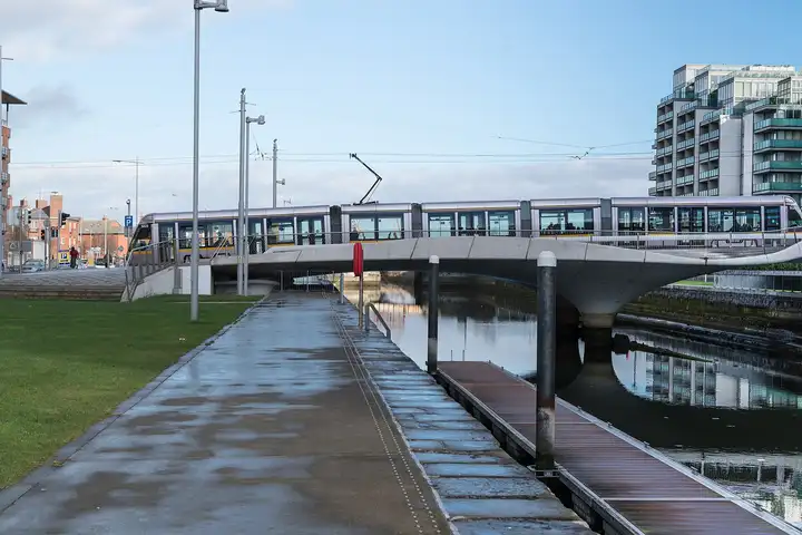 Eine Luas-Straßenbahn fährt über die flache Betonbrücke am Spencer Dock, daneben ein nasser Kaiweg und ein Schwimmsteg, rechts ein Wohnblock.