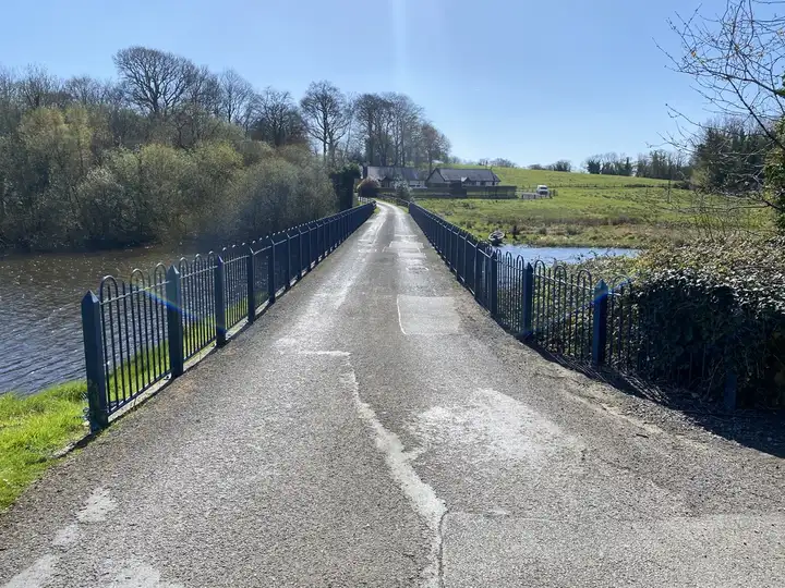 A narrow road bridge with blue railings crosses the water to Belle Isle, with estate buildings and trees on the far side.
