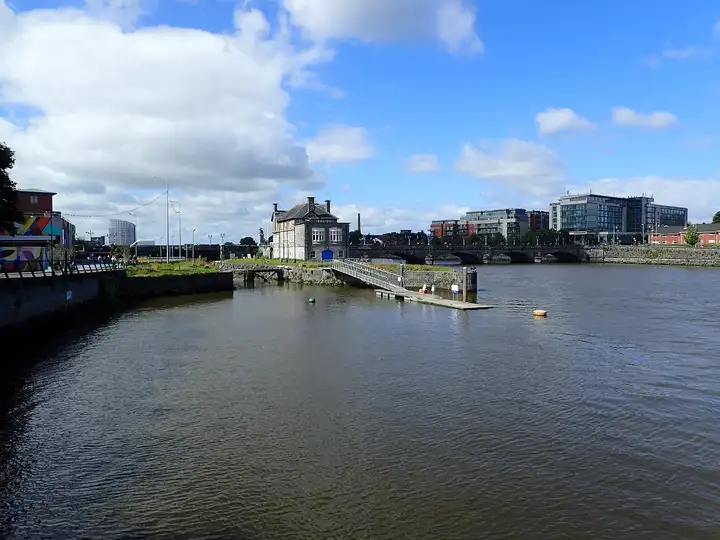 Der Shannon fließt in Limerick an einem alten Steinhaus mit einer Gangway hinab zu einem Ponton vorbei, dahinter die Sarsfield Bridge und moderne Gebäude der Stadt.