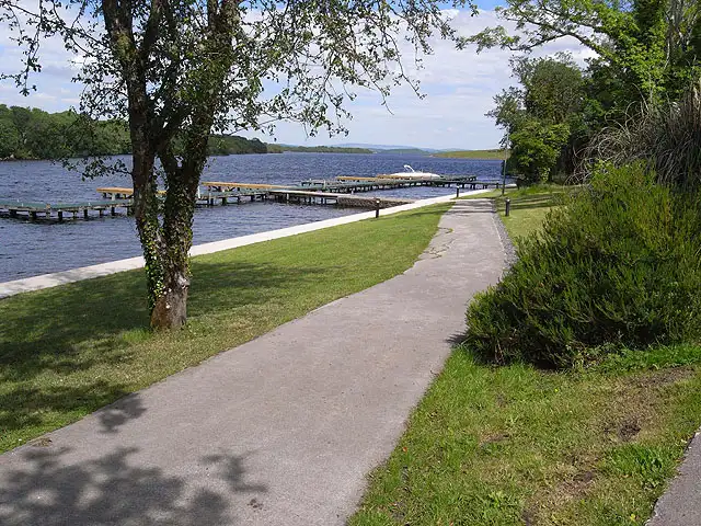 A paved path under a leafy tree runs along the shore of Lusty Beg to its timber jetties, where a cruiser is moored on the blue lough.