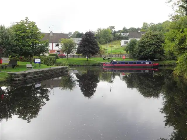 A red narrowboat moored on the still canal at Ballinamore, with trees, houses and a stone-edged quay reflected in the water.