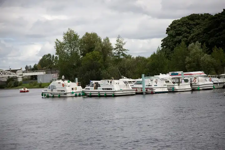 A row of white hire cruisers moored along a jetty on the Shannon in Athlone, with trees behind and the white railway bridge on the left.