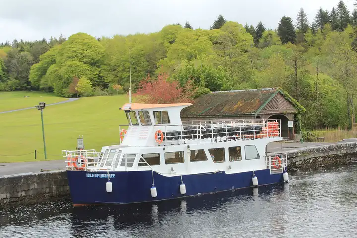 The blue-and-white cruise boat Isle of Inisfree moored at the stone pier of Ashford Castle near Cong, in front of a moss-roofed boathouse, lawns and spring woodland.