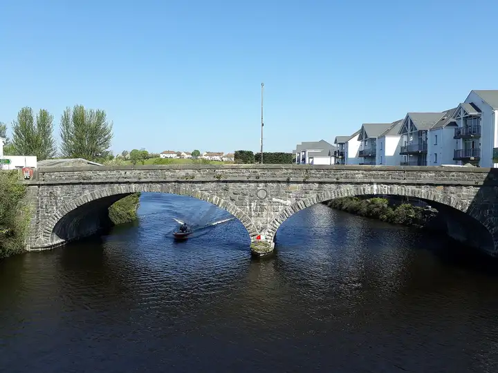 A small motorboat passes under a two-arched stone bridge over the River Erne in Enniskillen, with modern riverside apartments on the right.