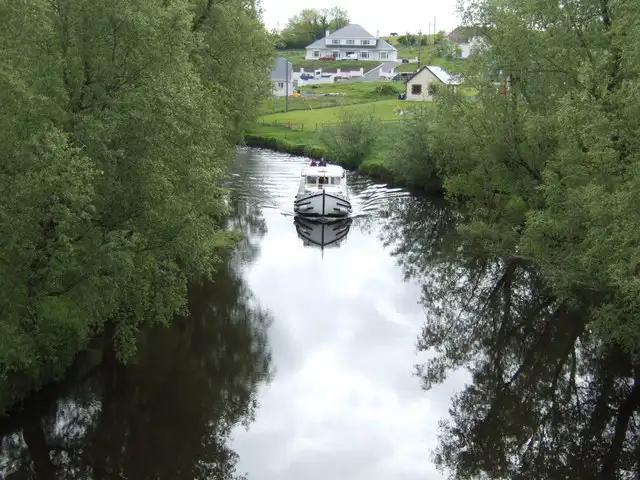 A cruiser on the tree-lined Woodford River at Aghalane, with the banks reflected in the calm water and houses on the slope behind.