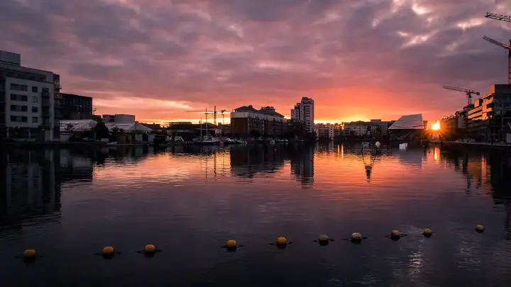 Coucher de soleil sur l’eau calme du Grand Canal Dock, avec une ligne de bouées jaunes au premier plan et les bâtiments et grues des quais en silhouette sur un ciel orangé.