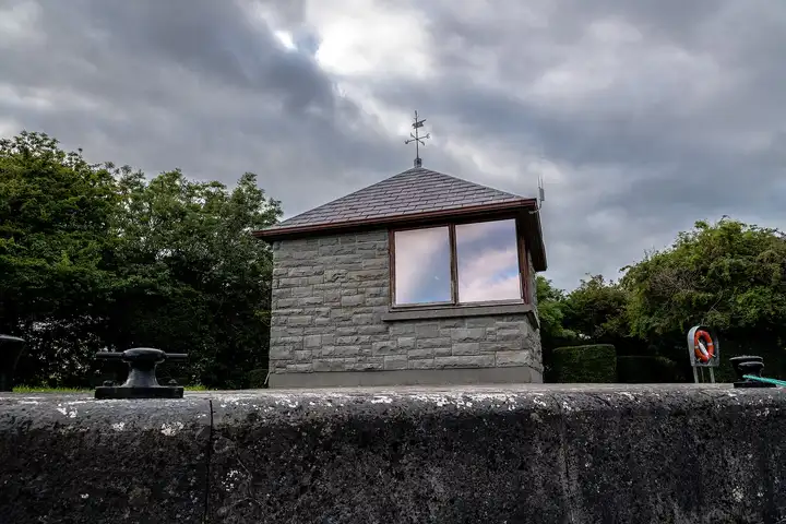 A small stone hut with a weather vane at Victoria Lock, behind the lock wall with its bollards and a lifebuoy, under a dark sky.