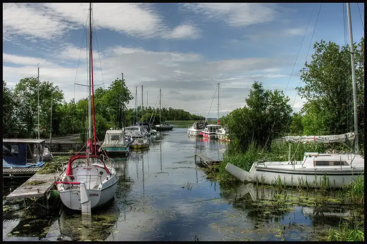 Een smalle havengeul bij Kilgarvan Quay met aangemeerde zeil- en motorboten, kleine houten steigers en waterlelies, die uitkomt op het open meer.