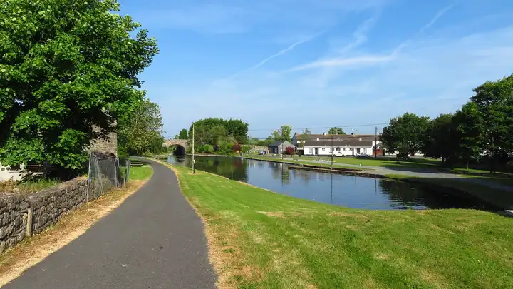 The greenway follows the canal into Ballynacargy, where the harbour basin reflects a blue sky beside lawns, a row of white cottages and a stone bridge.