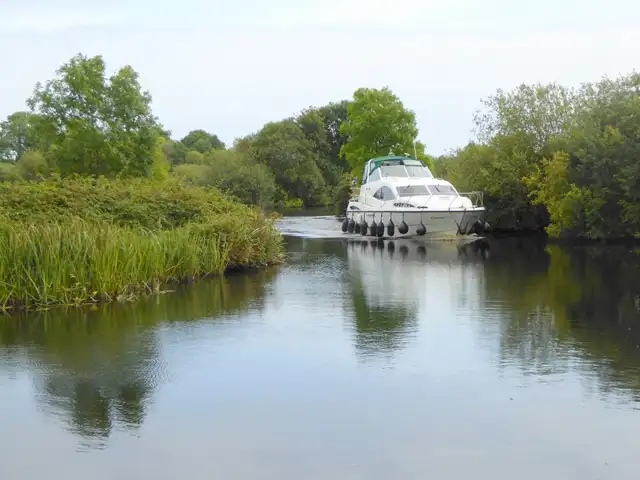 Ein weißes Kajütboot auf dem gewundenen, schilfgesäumten Scarriff River bei Tuamgraney Quay zwischen grünen Bäumen.