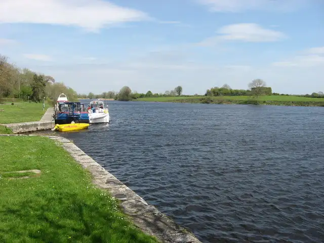 Motorboten, een aak en een klein geel bootje afgemeerd aan het eind van Meelick Quay op de oever van Galway, met daarachter de brede Shannon.