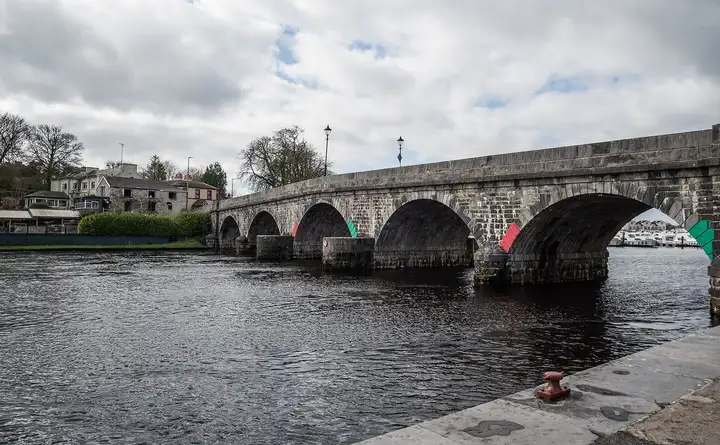 De stenen bogen van de brug van Carrick-on-Shannon, gemarkeerd met rode en groene vaarwegborden, rijzen op uit de donkere rivier naast een kade met een rode bolder.