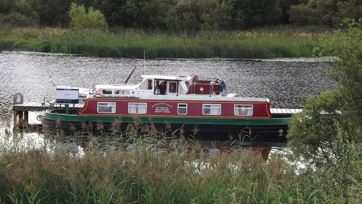 Een rood-groene kanaalboot ligt aangemeerd aan een steiger tussen het riet bij Crom aan Upper Lough Erne.