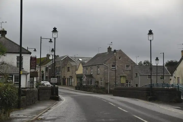 The A35 crosses the Kesh River on a low stone-walled bridge into Kesh village, with grey stone houses and lamp posts under a cloudy sky.