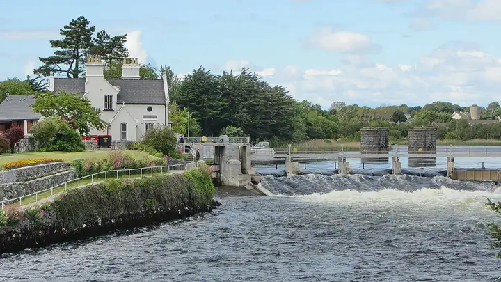 Schuimend water stroomt door de schuiven van de zalmstuw in de River Corrib in Galway, naast een wit huis en bloembedden op de oever.