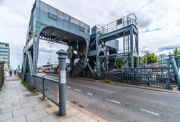 Die grauen, genieteten Stahlträger und gebogenen Rollsegmente einer Scherzer-Rollklappbrücke stehen neben Straße und Gehweg am Liffey-Ende des Spencer Dock.