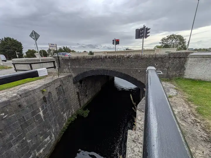 De stenen kolk van de 12e sluis onder de boog van de verkeersbrug, met een zwart-witte balk op de voorgrond en verkeerslichten op de brug.