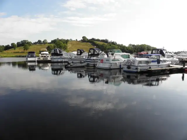 A row of motor cruisers moored at Bellanaleck is mirrored in the still water of the River Erne, with a green hillside behind.