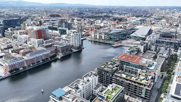Aerial view of Grand Canal Dock, a long basin of water lined with brick apartment blocks and modern offices, with the city stretching to the hills on the horizon.