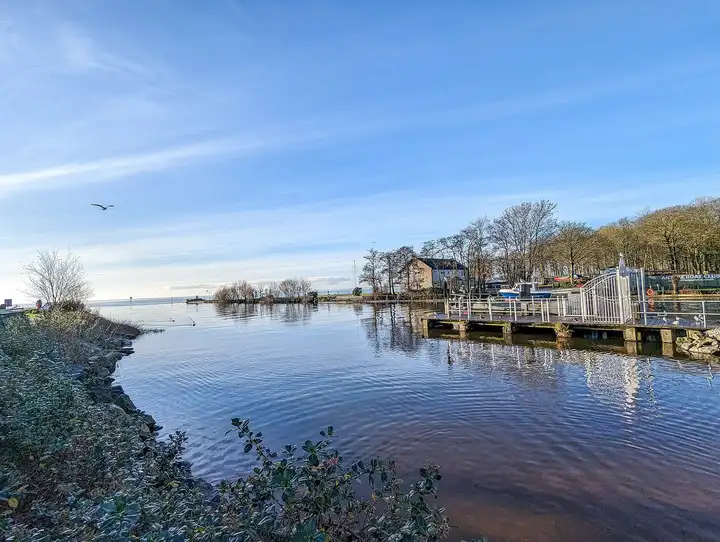 Calm water at Antrim’s lough shore, with the jetty and white railings of Antrim Boat Club on the right and Lough Neagh opening out beyond.