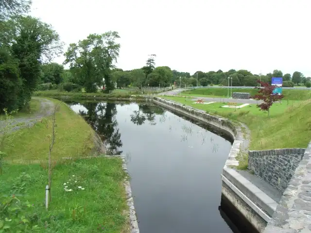 Seen from a bridge, the stone-edged channel of Mosstown Harbour near Keenagh curves between grassy banks and trees towards a lawn and a road.