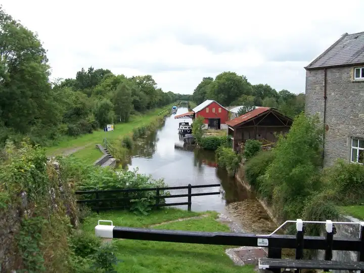 Het kanaal voorbij de 33e sluis bij Belmont, met een rode schuur en oude botenhuizen bij een kleine haven met afgemeerde motorboten, gezien langs een zwart-witte sluisbalk.