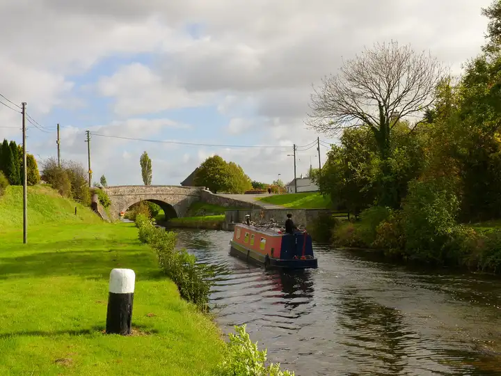 A red-and-blue narrowboat cruising along the canal at Daingean towards a stone road bridge, with a black-and-white bollard on the bright green towpath.