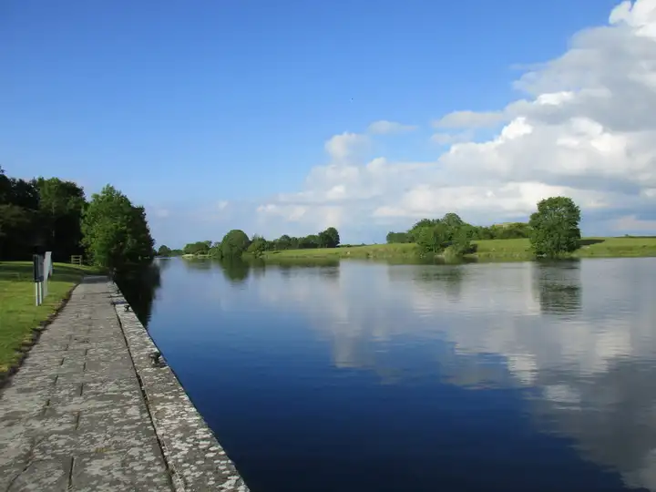 The long stone quay at Meelick runs beside the calm Shannon, which mirrors a blue sky and white clouds above flat green fields.