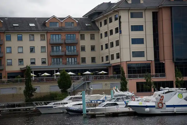 Motor cruisers moored at the floating pontoons of the marina in the centre of Athlone, in front of a large riverside hotel with a terrace.