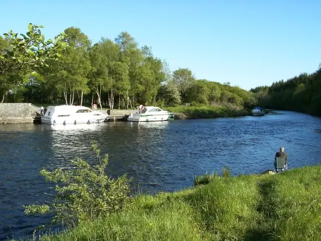 Two hire cruisers moored along the bank of the Shannon at Battlebridge on a sunny day, with an angler sitting on the grassy near bank.