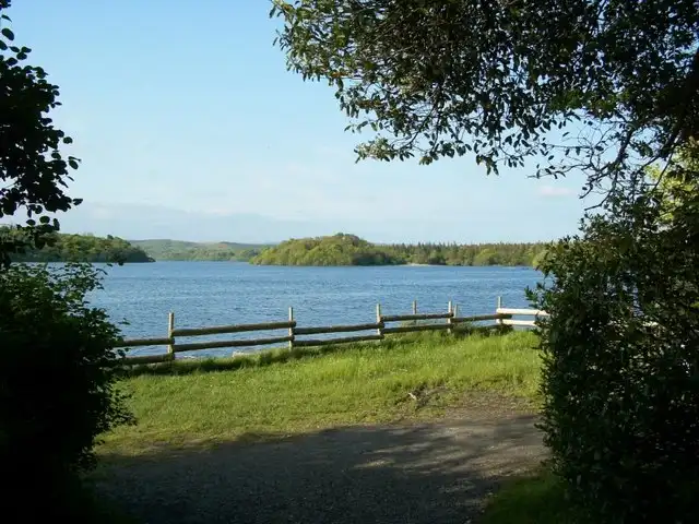 Lough Key en een bebost eiland, gezien door een opening tussen de bomen aan de oever van het bospark, achter een laag houten hek.