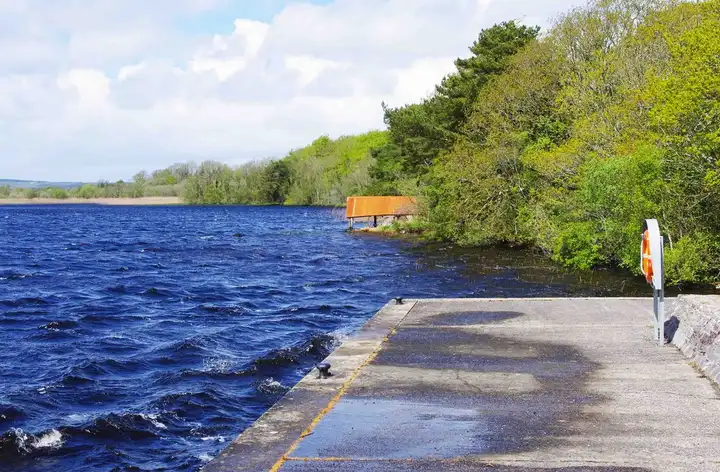 The concrete quay at Rossmore beside choppy blue water, with an old boathouse among the trees along the wooded shore.