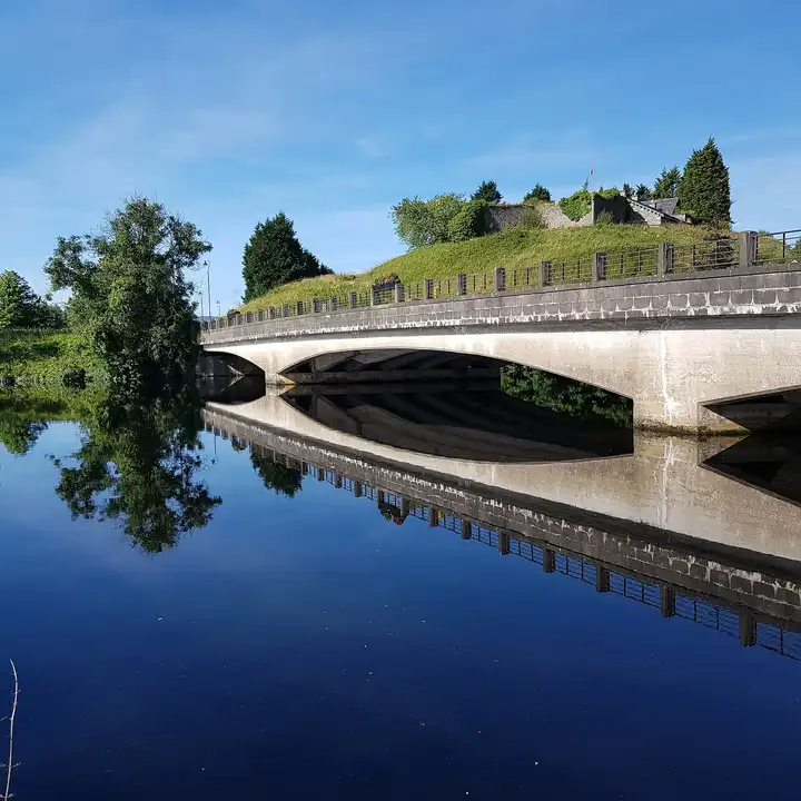 Under a deep-blue sky, the pale arches of Belleek Bridge and the grassy hill above it are mirrored in the still River Erne.