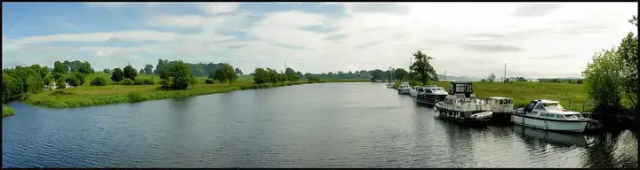 A wide view along the Shannon from Portumna Bridge, with green meadows on the left and a row of cruisers moored on the right bank.