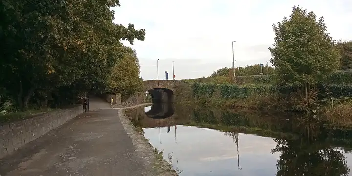 The stone arch of Mullen Bridge is mirrored in the calm canal on an overcast day, with the towpath running beneath trees on the left.