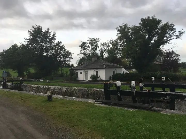 Een gerestaureerd wit sluiswachtershuisje staat achter de stenen muren en deuren van sluis 44 bij Killashee, omlijst door grote bomen onder een bewolkte lucht.