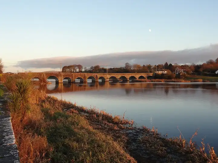 Low sun lights the long stone bridge at O’Briensbridge and its reflection in the calm Shannon, with reeds along the near bank.