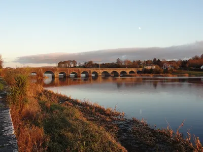 Low sun lights the long stone bridge at O’Briensbridge and its reflection in the calm Shannon, with reeds along the near bank.
