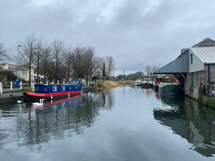 Boote liegen an beiden Seiten des Hafens von Naas, links ein blaues Kanalboot und Schwäne, rechts ein Vordach über dem Kai an den Steinspeichern.