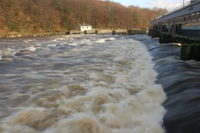 Braunes Hochwasser stürzt unterhalb der Hochwassertore an den Cutts über die Schwelle, rechts das Torbauwerk, dahinter herbstlicher Wald.