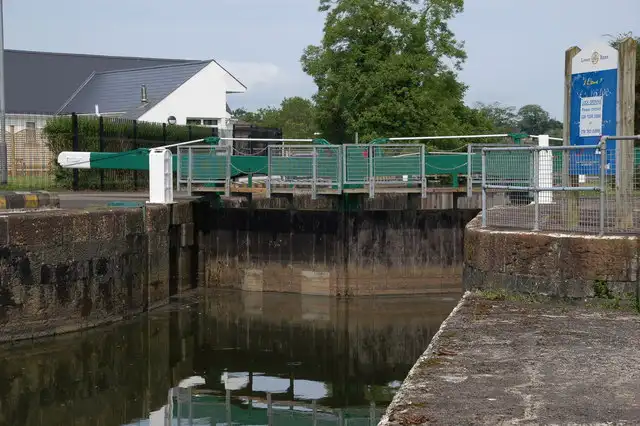 Die Tore der Schleuse an den Cutts mit grünen Geländern am Laufsteg, einem Schild neben der Schleuse und einem Haus hinter den Bäumen.